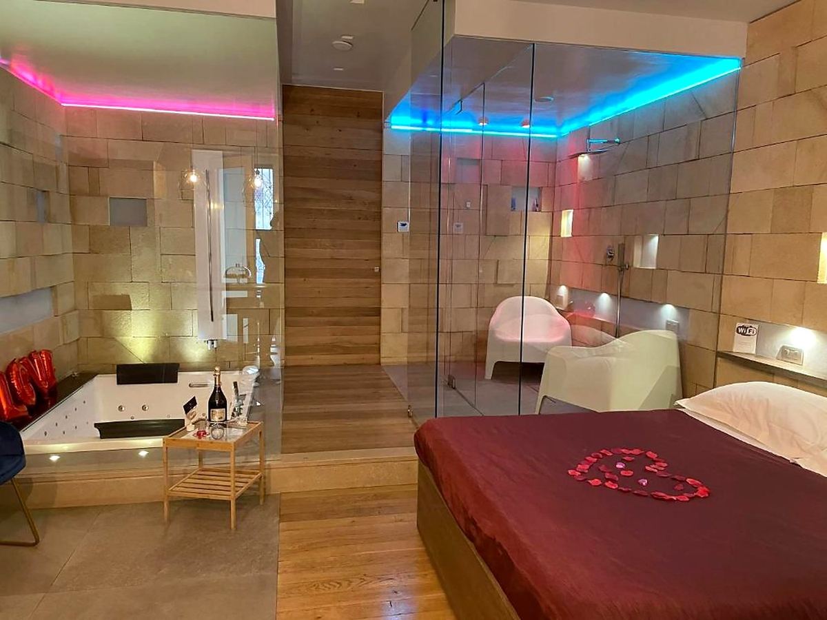 La suite préparée pour le forfait romantique : pétales en cœur sur le lit bordeaux, baignoire balnéo et lumières colorées